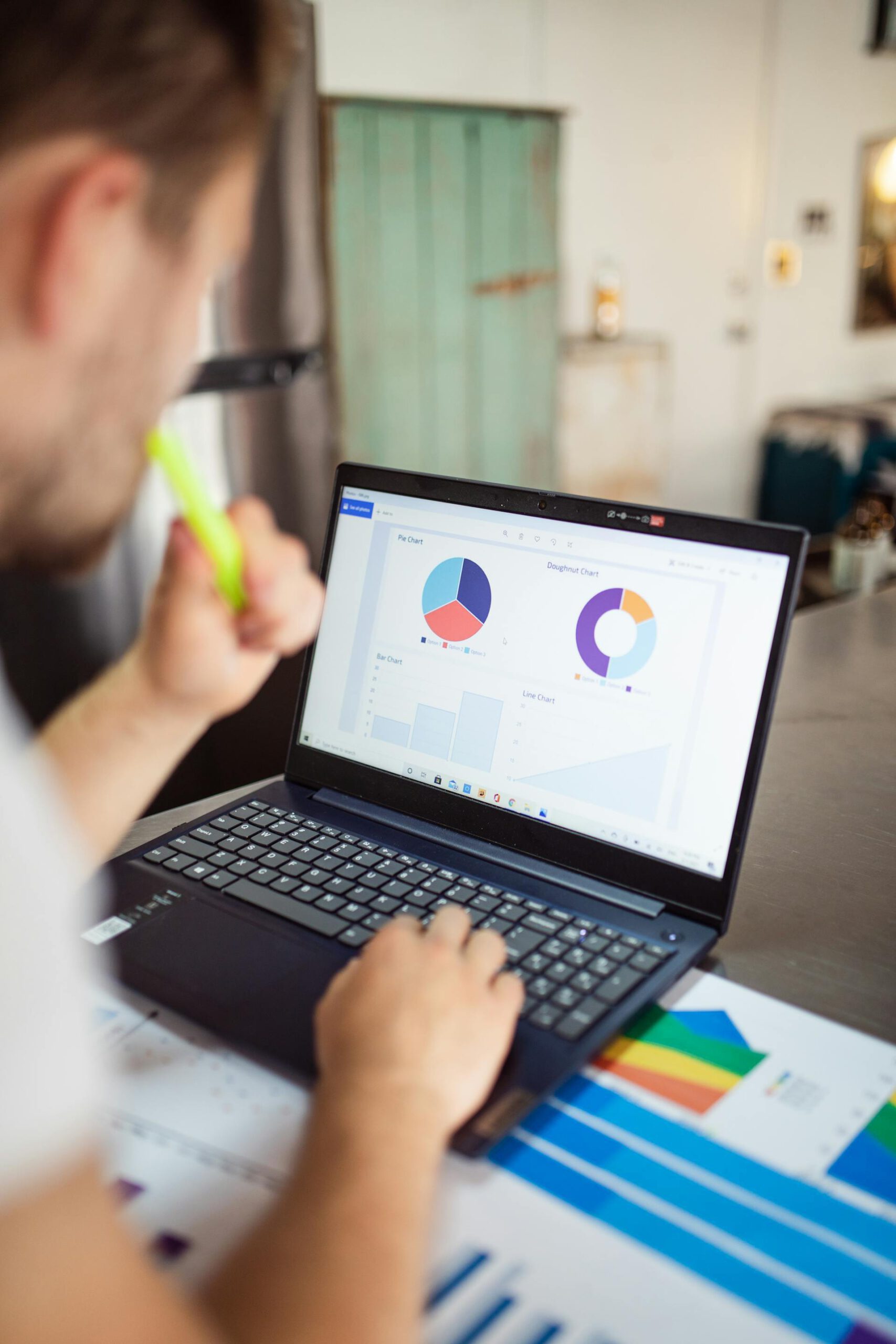 Person working on a laptop displaying various charts, including pie, doughnut, bar, and line charts, with printed graphs on the desk.